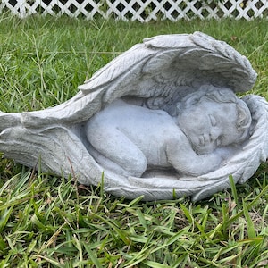 May include: A grey concrete statue of a sleeping angel with wings. The angel is lying on its side with its head resting on its arm. The wings are spread out behind the angel.