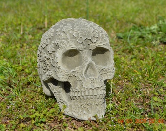 Puede incluir: Una escultura de calavera de piedra gris con intrincados diseños florales tallados en la superficie. La calavera está sentada sobre un lecho de hierba verde.