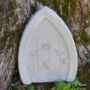 May include: White concrete fairy door with a heart, two butterflies, and a crown design. The door is attached to a tree trunk.