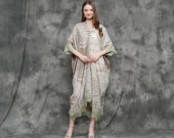 May include: A woman wearing a long, flowing, beige and white patterned dress with lace trim. The dress has a loose, flowing silhouette and a high neckline. The woman is standing in front of a gray textured background.