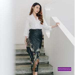 May include: A woman wearing a white long-sleeved blouse with a black and gold patterned wrap skirt. She is standing on a staircase and holding a small clutch.