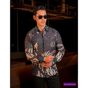 Puede incluir: Una camisa de manga larga con un estampado batik en tonos morado, verde azulado y crema. La camisa presenta un diseño floral y de hojas. La imagen también muestra a una persona con gafas de sol.