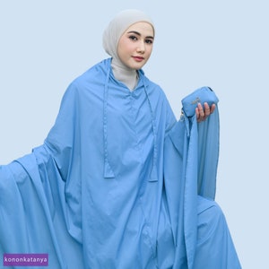 May include: A light blue prayer garment with a matching small pouch. The garment has a hood with drawstrings and a flowing design. The pouch is held in the hand and has a gold-coloured logo. The background is a light blue colour.