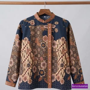 Könnte beinhalten: Ein marineblaues Langarmhemd mit Stehkragen und einem braunen und beigefarbenen Blumen-Batikmuster. Das Hemd hat einen vertikalen braunen Streifen auf der Vorderseite und hängt an einem Holzbügel.
