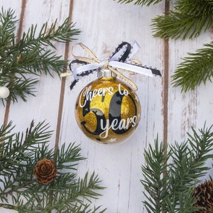 May include: A gold glitter Christmas ornament with the text "Cheers to 50 years" in white script. The ornament has a black number 50 and is adorned with a black and white ribbon and gold ribbon. The ornament is surrounded by pine branches.
