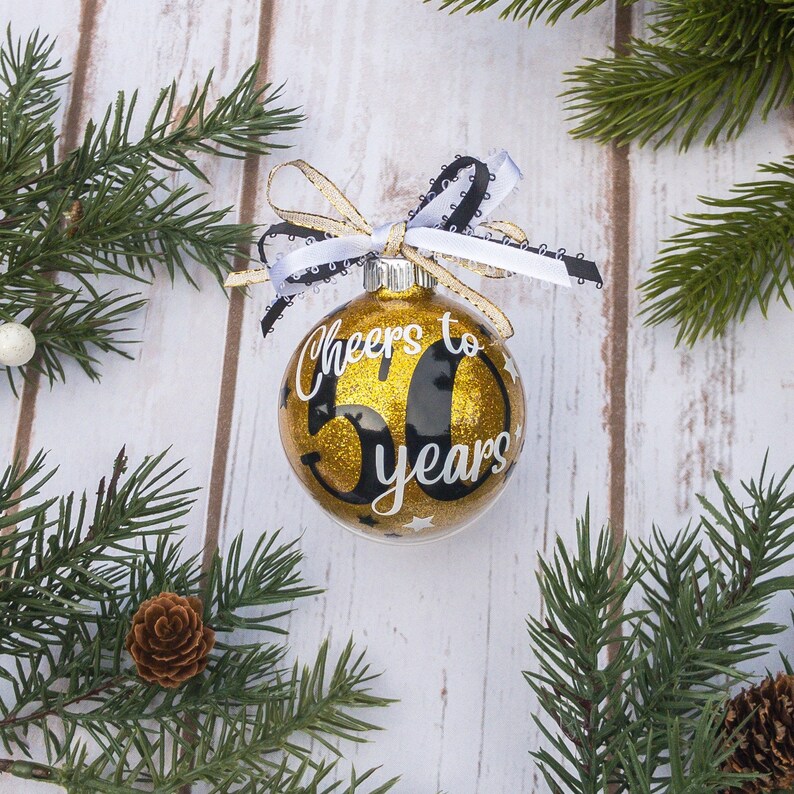 May include: A gold glitter Christmas ornament with the text "Cheers to 50 years" in white script. The ornament has a black number 50 and is adorned with a black and white ribbon and gold ribbon. The ornament is surrounded by pine branches.