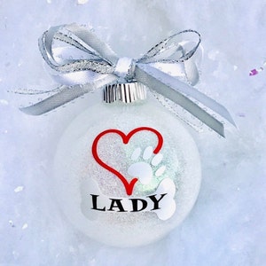 May include: White glitter Christmas ornament with a red heart and a white paw print inside the heart. The ornament also has a white bone with the name "LADY" written on it.