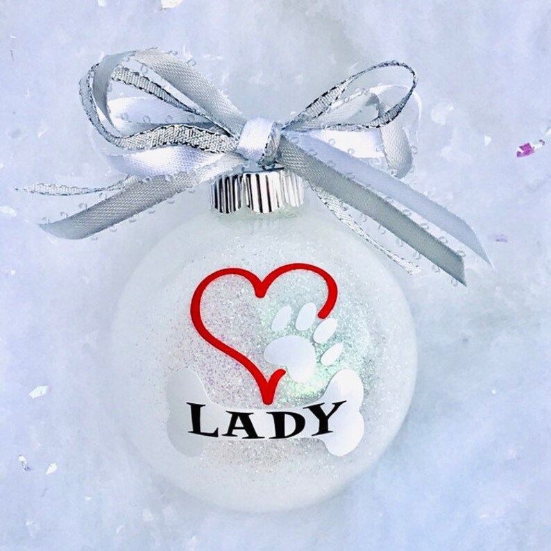 May include: White glitter Christmas ornament with a red heart and a white paw print inside the heart. The ornament also has a white bone with the name "LADY" written on it.