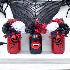 May include: Three Halloween-themed mason jars. Two are red with black bat silhouettes, and one is black with a vampire mouth graphic. Each jar is decorated with black, red, and white paper flowers and tied with a black ribbon.