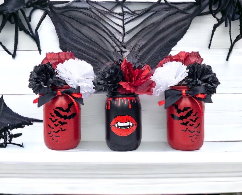 May include: Three Halloween-themed mason jars. Two are red with black bat silhouettes, and one is black with a vampire mouth graphic. Each jar is decorated with black, red, and white paper flowers and tied with a black ribbon.