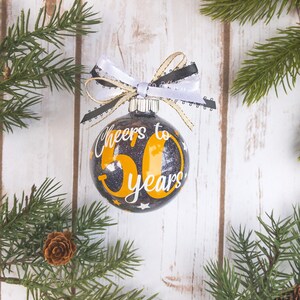 May include: A black and gold glitter Christmas ornament with the text "Cheers to 50 years" in white and gold. The ornament is decorated with a white, black, and gold ribbon. The background is a white wooden surface with pine branches.