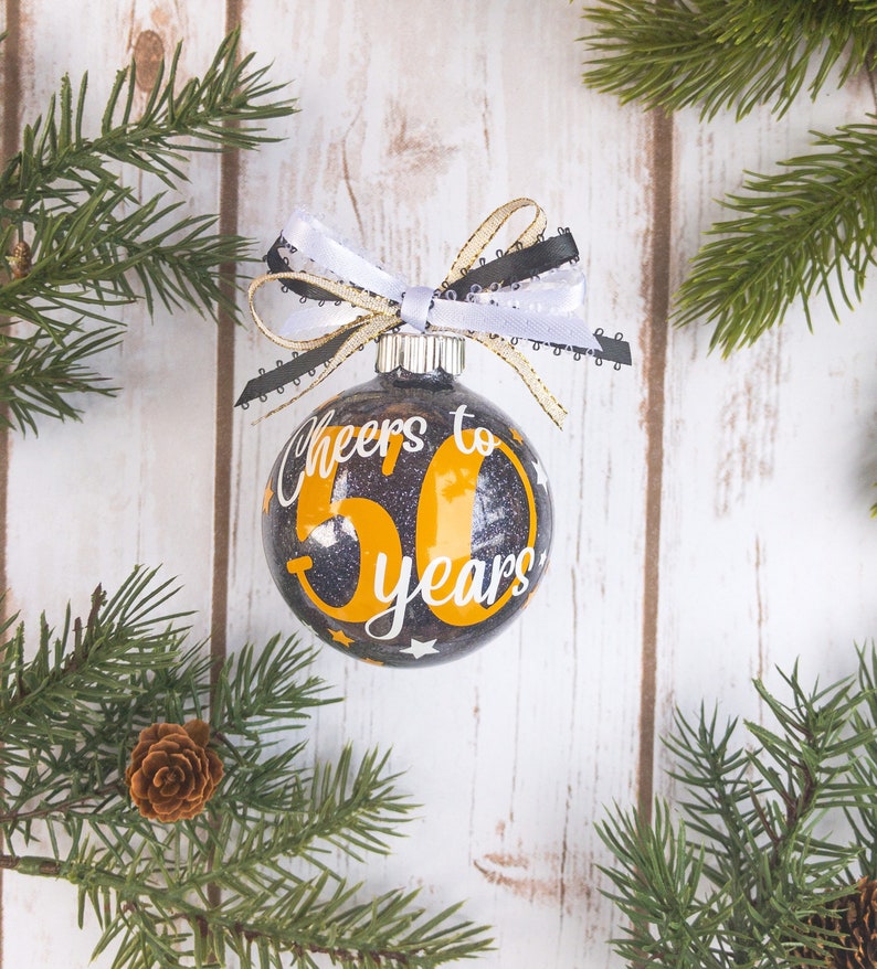 May include: A black and gold glitter Christmas ornament with the text "Cheers to 50 years" in white and gold. The ornament is decorated with a white, black, and gold ribbon. The background is a white wooden surface with pine branches.