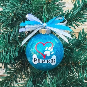 May include: A blue glitter Christmas ornament with a red heart and a white paw print. The ornament has a white bone with the name "PIPER" written on it.
