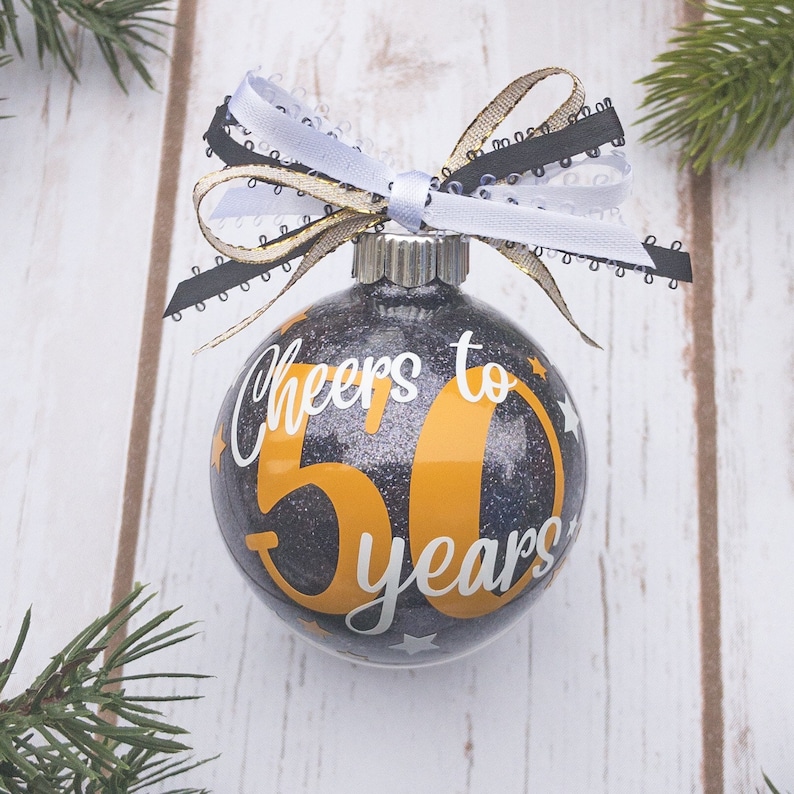 May include: Glittery black and silver Christmas ornament with the text "Cheers to 50 years" in white and gold. The ornament is topped with a decorative bow in black, white, and gold. Festive holiday decor.