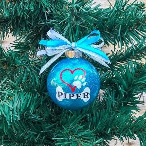 May include: A blue glitter Christmas ornament with a white bone, red heart, and paw print. The ornament is personalized with the name "PIPER".