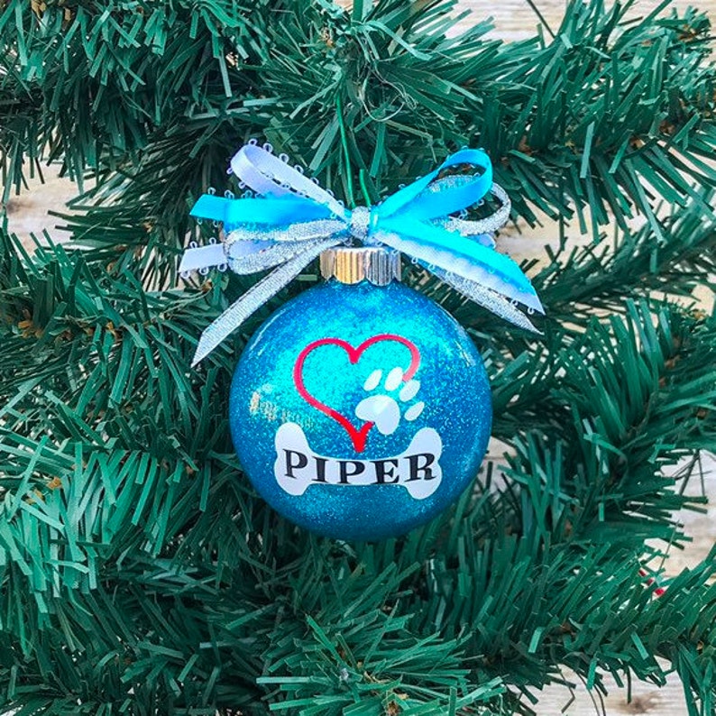 May include: A blue glitter Christmas ornament with a white bone, red heart, and paw print. The ornament is personalized with the name "PIPER".