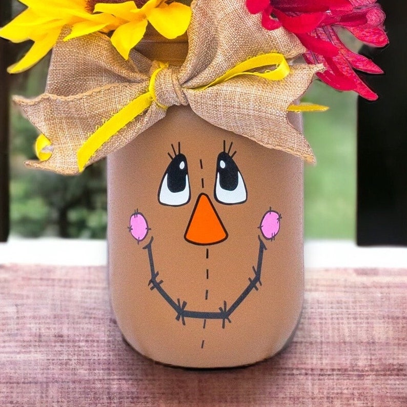 Peut inclure: Un bocal en verre brun avec un visage de  &eacute;pouvantail peint. Le bocal est d&eacute;cor&eacute; d'un n&oelig;ud en toile de jute et de tournesols jaunes.