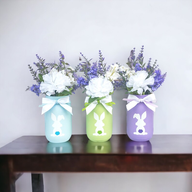 May include: Three mason jars painted in pastel blue, green, and purple with a white bunny silhouette on each. Each jar is filled with purple and white flowers.