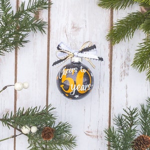 May include: A black and gold Christmas ornament with the text "Cheers to 50 years". The ornament has a white and black ribbon bow at the top. The background is a white wooden surface with pine branches.
