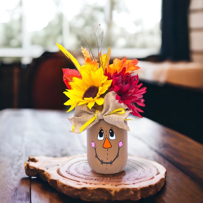 Peut inclure: Un bocal en verre peint avec un visage de l'&eacute;pouvantail et un arrangement floral d'automne. Les fleurs sont jaunes, rouges et orange.
