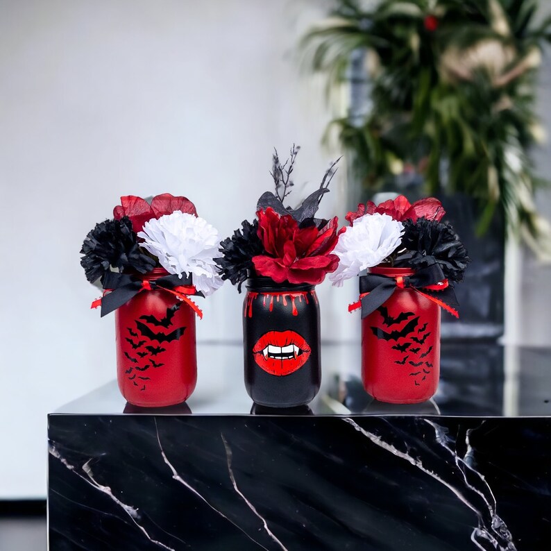 May include: Three Halloween-themed mason jars with floral arrangements. Two jars are red with bat silhouettes, and one is black with a vampire mouth design. Each jar holds a mix of red, black, and white flowers, tied with black ribbons.