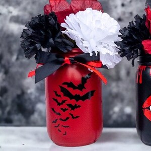 May include: A red painted mason jar decorated with black bat silhouettes, black ribbon, and red and white paper flowers. The jar is part of a Halloween-themed arrangement.