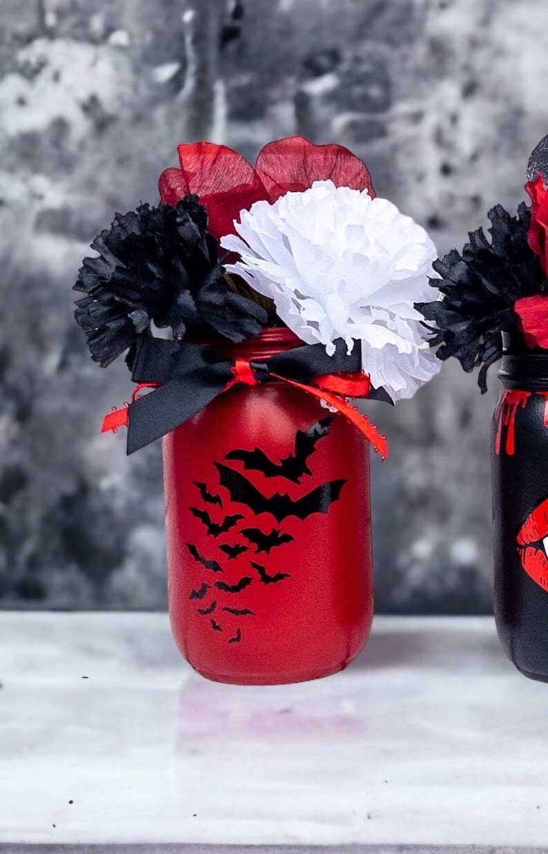 May include: A red painted mason jar decorated with black bat silhouettes, black ribbon, and red and white paper flowers. The jar is part of a Halloween-themed arrangement.