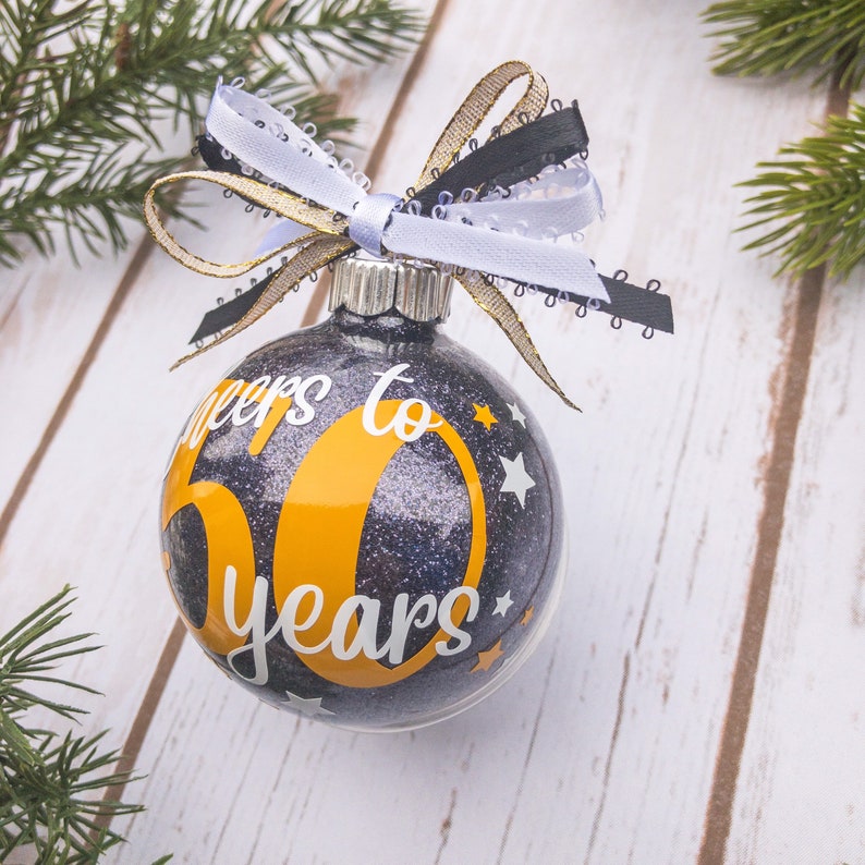 May include: Glitter-filled Christmas ornament with the text "Cheers to 50 years" in white and orange. The ornament is topped with a decorative bow of white, black, and gold ribbons. The background is a white wooden surface with pine branches.
