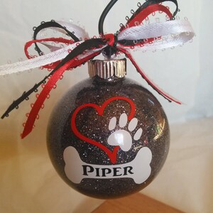 May include: A black glitter Christmas ornament with a red heart, a white paw print, and a white bone with the name "PIPER" on it. The ornament is decorated with a red, white, and black ribbon.