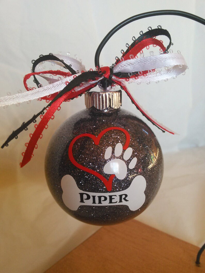 May include: A black glitter Christmas ornament with a red heart, a white paw print, and a white bone with the name "PIPER" on it. The ornament is decorated with a red, white, and black ribbon.