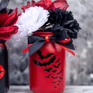 May include: A red painted mason jar with a bat silhouette design, filled with red, white, and black paper flowers. A black ribbon with red trim is tied around the jar. The jar is a decorative item.