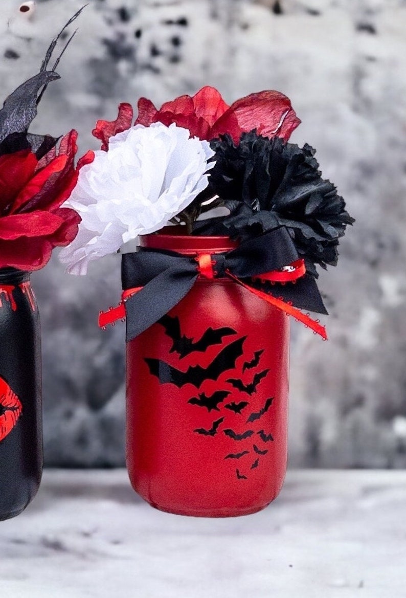 May include: A red painted mason jar with a bat silhouette design, filled with red, white, and black paper flowers. A black ribbon with red trim is tied around the jar. The jar is a decorative item.