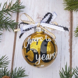 May include: A gold glitter Christmas ornament with the text "Cheers to 50 years" in white script. The ornament is decorated with small white stars and a black and white ribbon bow. Green pine branches surround the ornament.