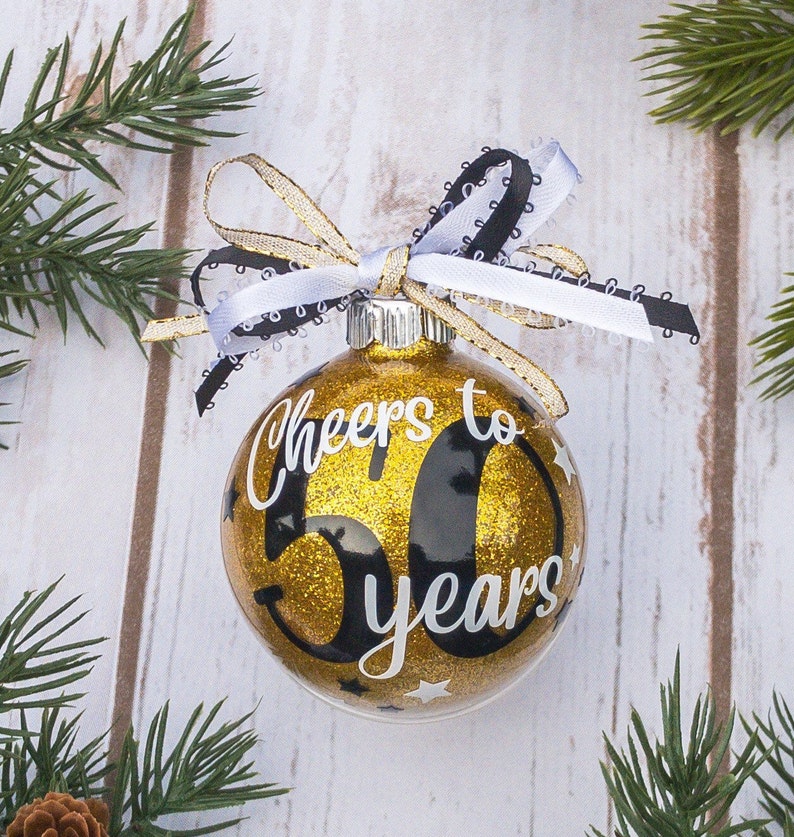 May include: A gold glitter Christmas ornament with the text "Cheers to 50 years" in white script. The ornament is decorated with small white stars and a black and white ribbon bow. Green pine branches surround the ornament.