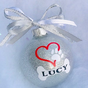 May include: Silver glitter Christmas ornament with a red heart and a white paw print. The ornament has a white bone with the name "Lucy" written on it.