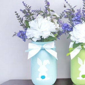 May include: Two mason jars painted in pastel blue and green with a white bunny silhouette. The jars are tied with white ribbon and filled with purple and white flowers.