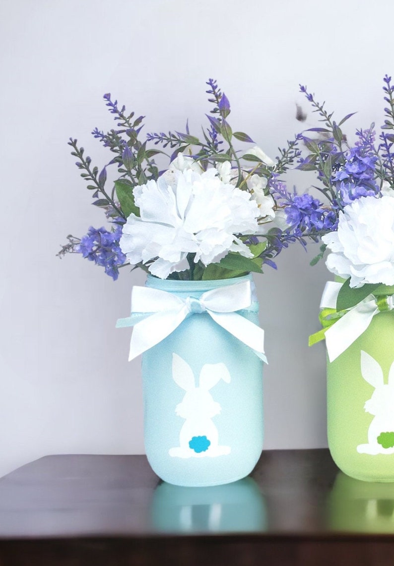 May include: Two mason jars painted in pastel blue and green with a white bunny silhouette. The jars are tied with white ribbon and filled with purple and white flowers.