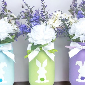 May include: Three mason jars painted in pastel blue, green, and purple with white bunny silhouettes. Each jar is decorated with a white ribbon and filled with a bouquet of white and purple flowers.