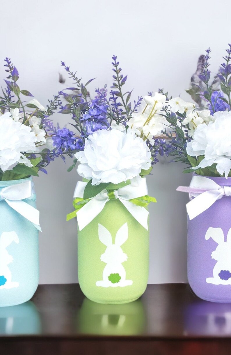 May include: Three mason jars painted in pastel blue, green, and purple with white bunny silhouettes. Each jar is decorated with a white ribbon and filled with a bouquet of white and purple flowers.