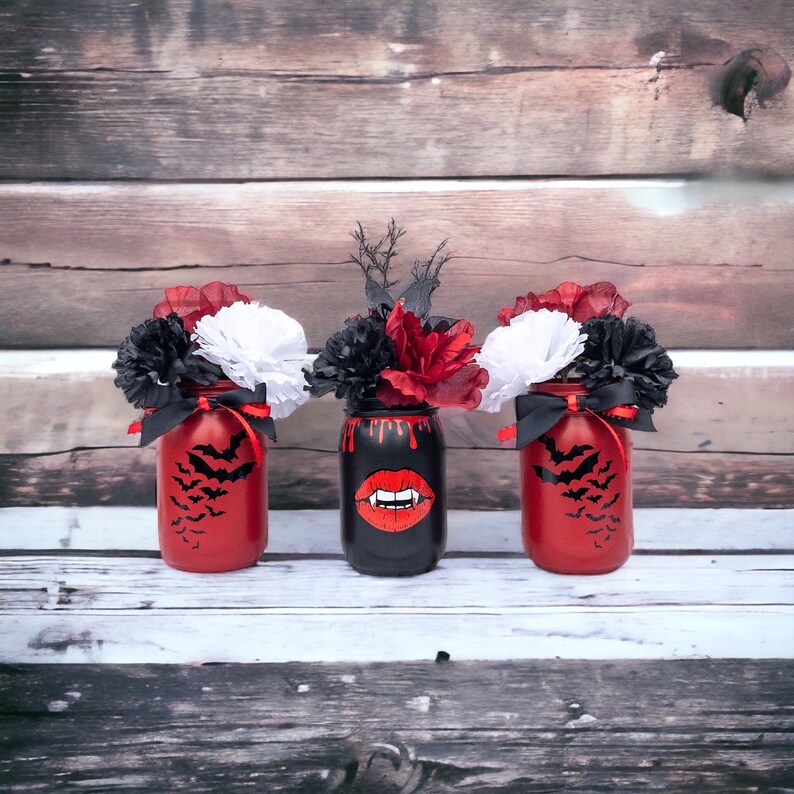 May include: Three Halloween-themed mason jars with floral arrangements. Two red jars feature black bat silhouettes, while the center black jar has a vampire mouth design. Each jar holds black, red, and white flowers, tied with black ribbons.