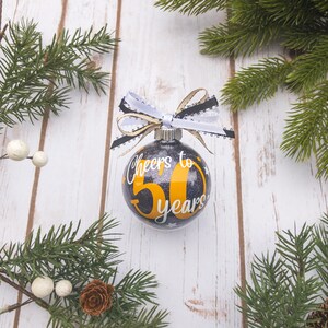 May include: A black and silver glitter Christmas ornament with the text "Cheers to 50 years" in white and yellow. The ornament is decorated with a white, black, and gold ribbon. The background includes pine branches and a white wooden surface.