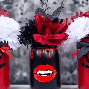 May include: Three Halloween-themed mason jars. One black jar features a red vampire mouth design and red dripping paint. The other two jars are red with black bat cutouts. Each jar holds a floral arrangement with red, white, and black flowers.