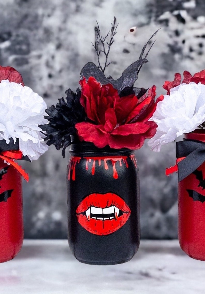 May include: Three Halloween-themed mason jars. One black jar features a red vampire mouth design and red dripping paint. The other two jars are red with black bat cutouts. Each jar holds a floral arrangement with red, white, and black flowers.