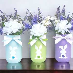 May include: Three mason jars painted in pastel colors, light blue, green, and purple, with white bunny silhouettes. Each jar is filled with a bouquet of white carnations and purple flowers.