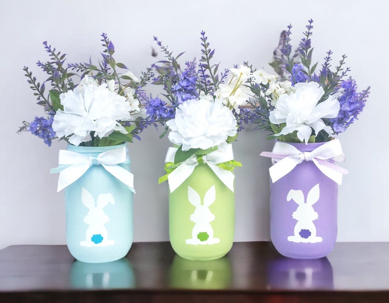 May include: Three mason jars painted in pastel colors, light blue, green, and purple, with white bunny silhouettes. Each jar is filled with a bouquet of white carnations and purple flowers.