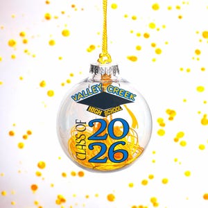 以下が含まれることがあります： 「Valley Creek High School Class of 2026」の文字が青と金で書かれた透明なガラス製のクリスマスオーナメント。オーナメントは金色の装飾材で満たされ、金色の紐で吊るされています。