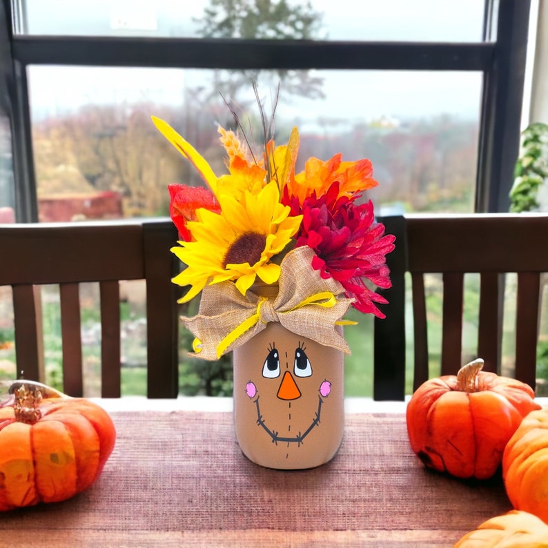Peut inclure: Un bocal en verre peint pour ressembler &agrave; un visage d'&eacute;pouvantail avec un n&oelig;ud en toile de jute et des fleurs d'automne &agrave; l'int&eacute;rieur. Le bocal est pos&eacute; sur une table en bois avec deux petites citrouilles orange.
