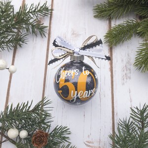 May include: A clear glass Christmas ornament with a black glitter background and the text "Cheers to 50 years" in white and gold. The ornament is decorated with a black, white, and gold ribbon bow and is surrounded by evergreen branches.