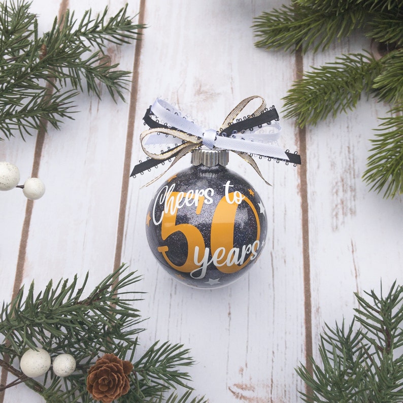 May include: A clear glass Christmas ornament with a black glitter background and the text "Cheers to 50 years" in white and gold. The ornament is decorated with a black, white, and gold ribbon bow and is surrounded by evergreen branches.