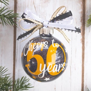 May include: Glittery black and gold Christmas ornament with the text "Cheers to 50 years" in white and gold. The ornament is decorated with a white, black, and gold ribbon bow. The ornament is surrounded by pine branches.